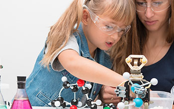 A woman teacher and girl collect molecules and conduct experiments. On the table are a robot and beakers with liquid inside.